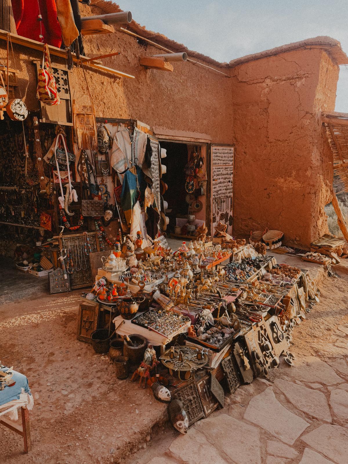 Souvenirladen in Ait Ben Haddou, umgeben von den typischen Lehmgebäuden des Dorfes. Das warme Sonnenlicht taucht die Szene in goldene Farben und hebt die kunstvoll handgefertigten Waren wie Teppiche und Schmuck hervor.
