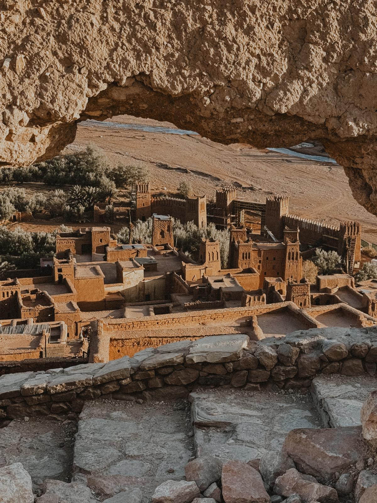 Luftaufnahme von Ait Ben Haddou, dem historischen Lehmdorf in Marokko, mit seinen charakteristischen roten Lehmgebäuden und der beeindruckenden Kasbah im Vordergrund, umgeben von der weiten Wüstenlandschaft und den Bergen des Atlasgebirges im Hintergrund.